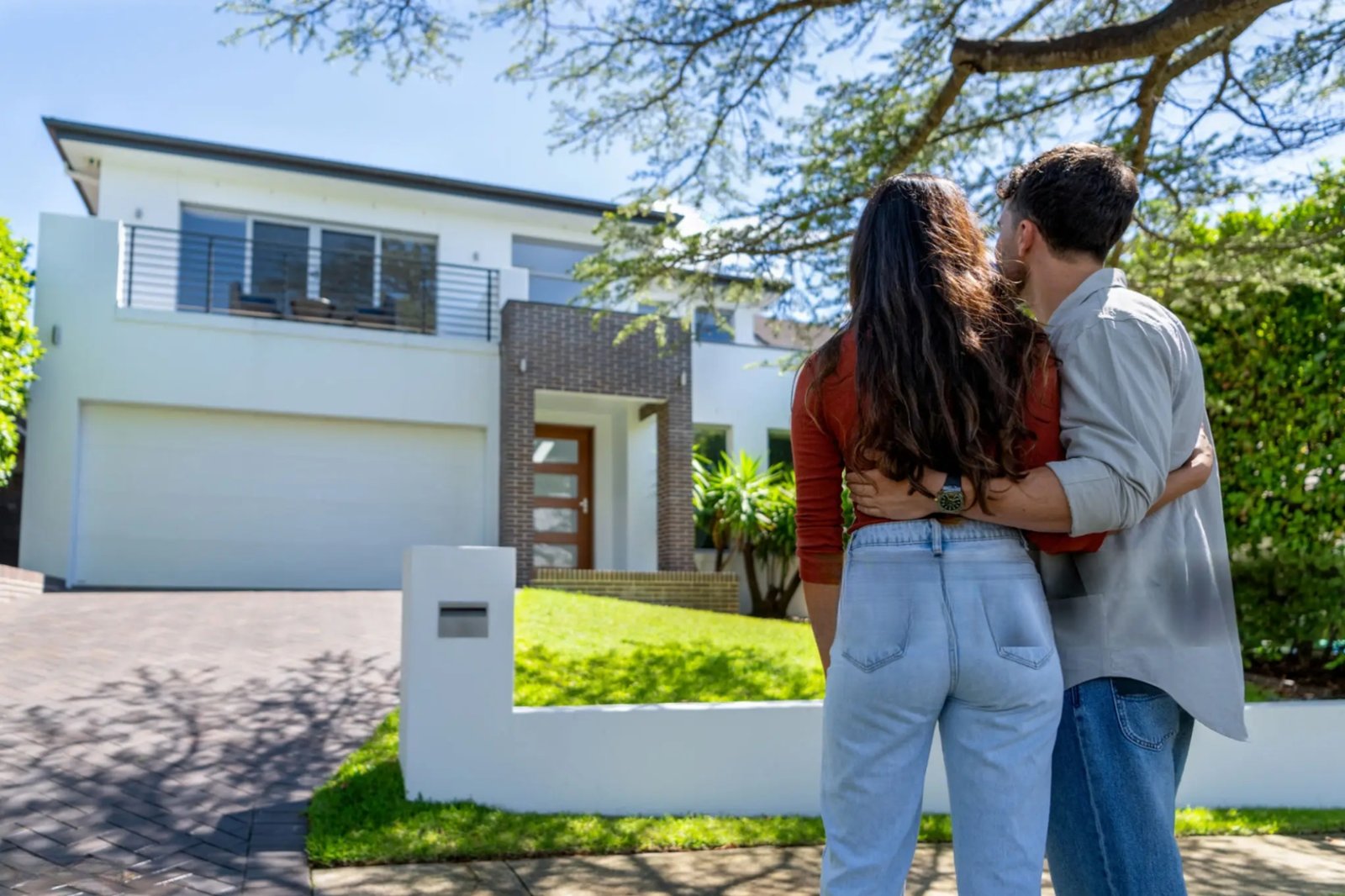 A young Australian couple standing in front of a house with their arms around each other, looking up at it together