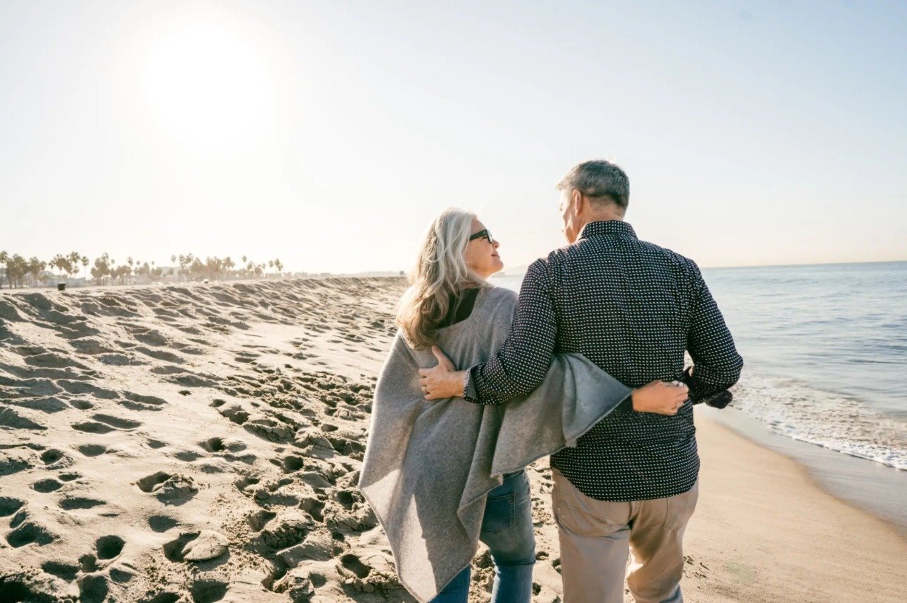A relaxed couple in their early 60s walking together along a beach path at sunset