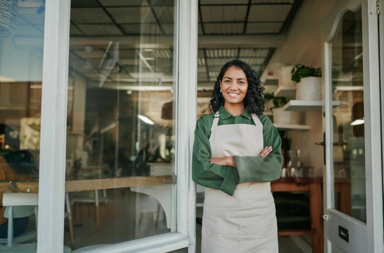A confident small business owner standing at the front entrance of her shop, arms crossed, smiling at the camera